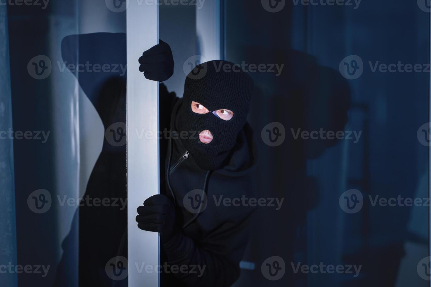 Criminal Concept. Masked burglar peeking into the house through an open glass door or window, copy space photo