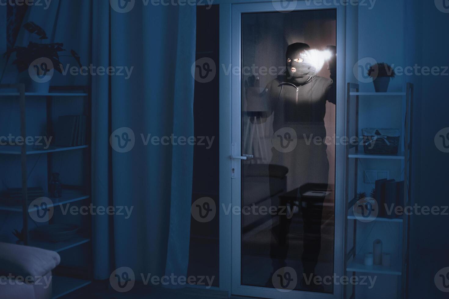 Housebreaking Concept. Masked thief looking into apartment through glass balcony door and using flashlight photo