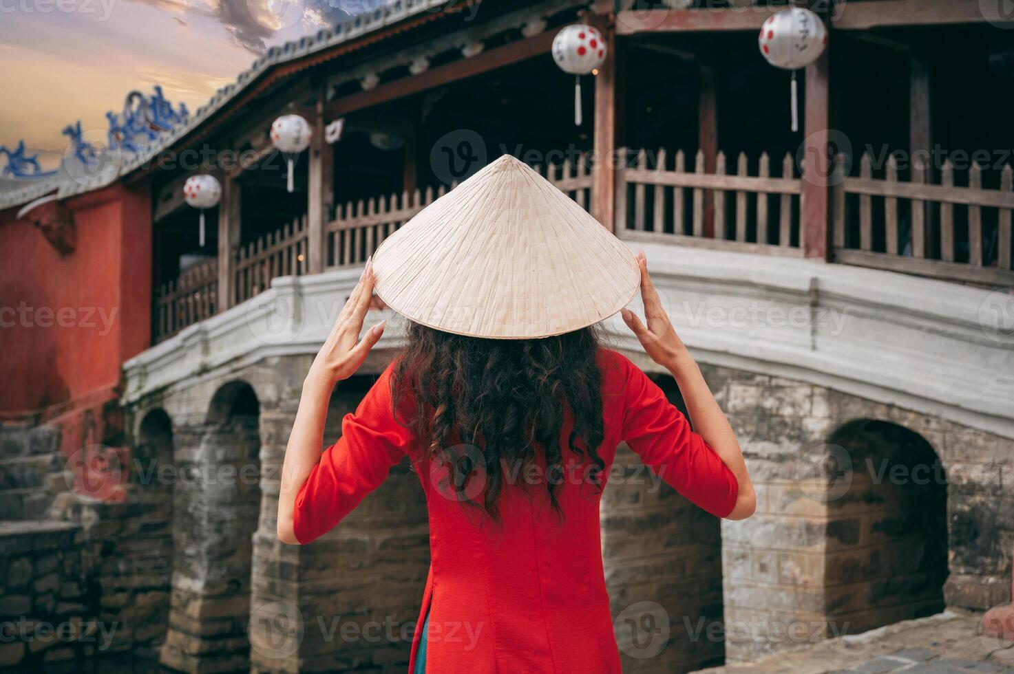 back of girl traveler tourist in Vietnamese hat at ancient Japanese covered bridge in old town in Hoi An in Vietnam photo