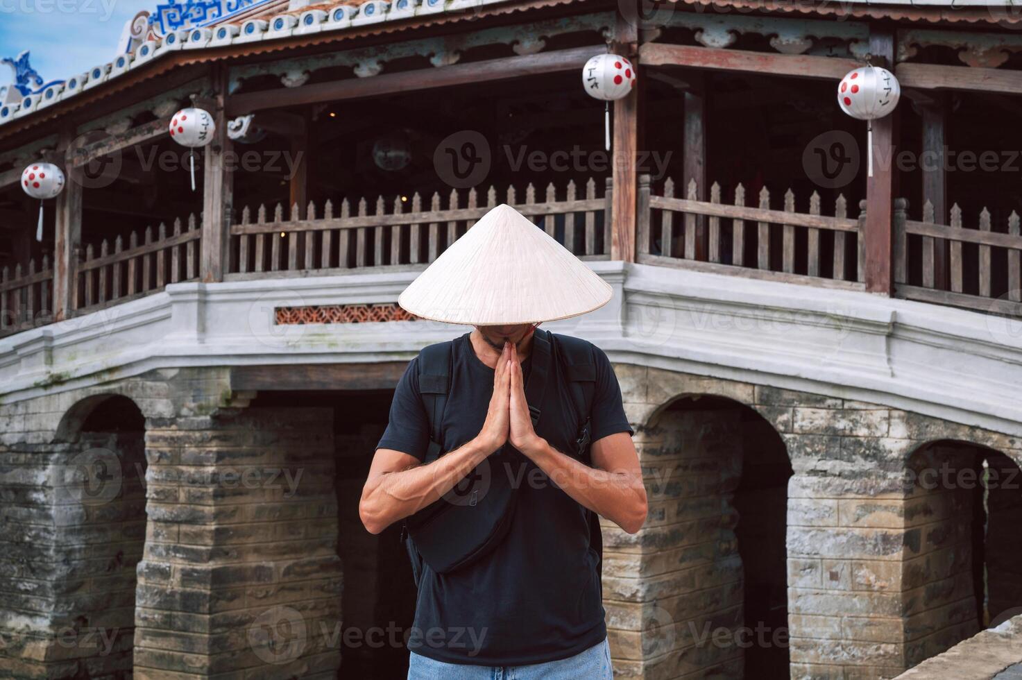 man traveler tourist in Vietnamese hat prays at the ancient Japanese covered bridge in old town in Hoi An in Vietnam in Asia photo