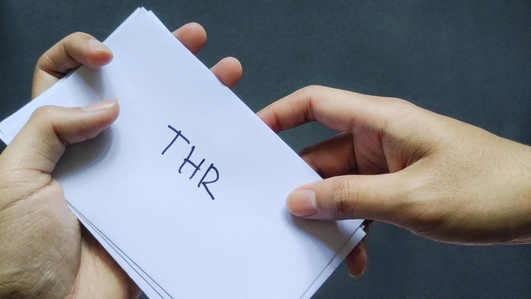 Woman holding a white envelope labeled THR or Hari Raya Allowance on a black background. Top view photo