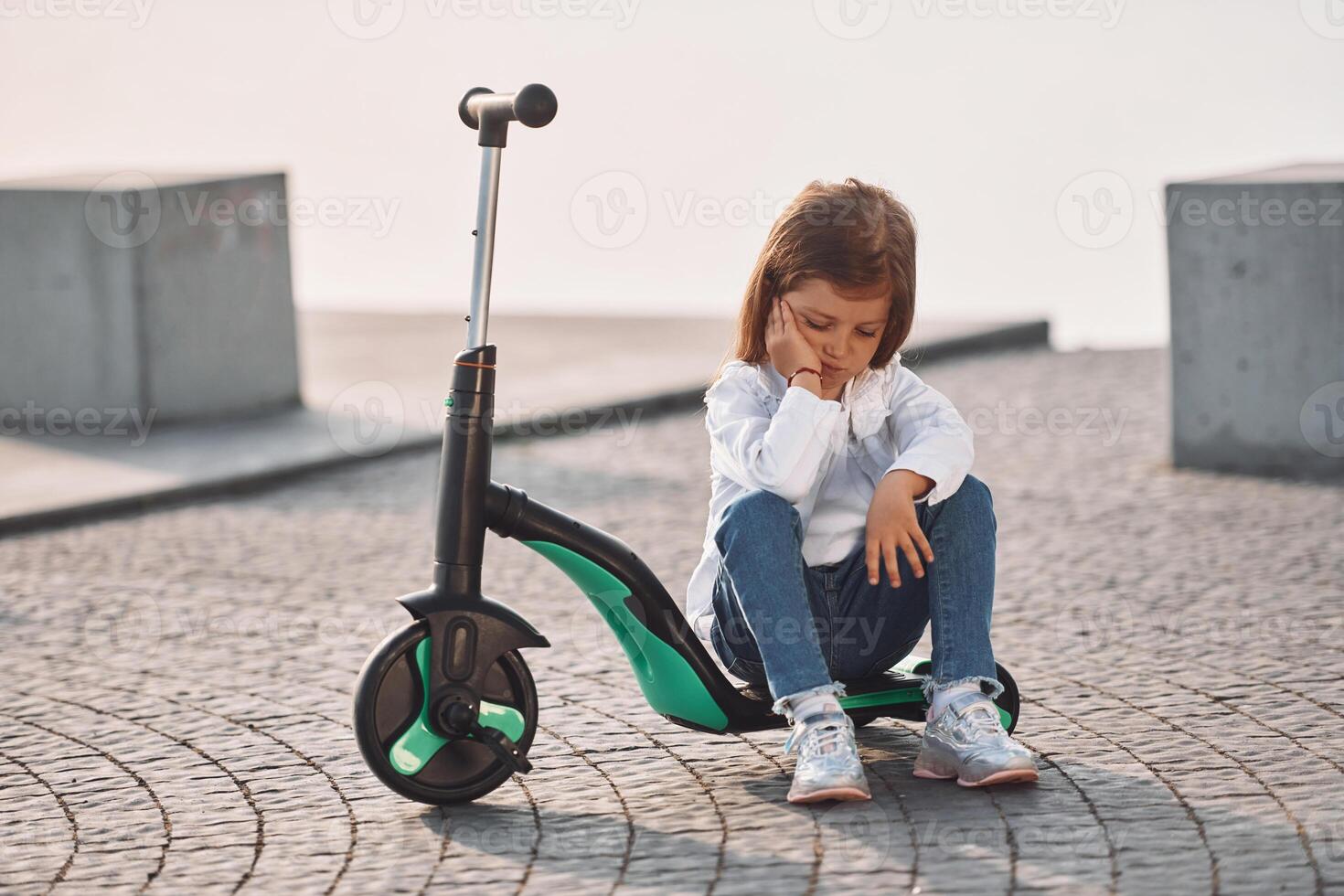 Sitting, bored. Cute little girl with scooter is outdoors in summertime, photo