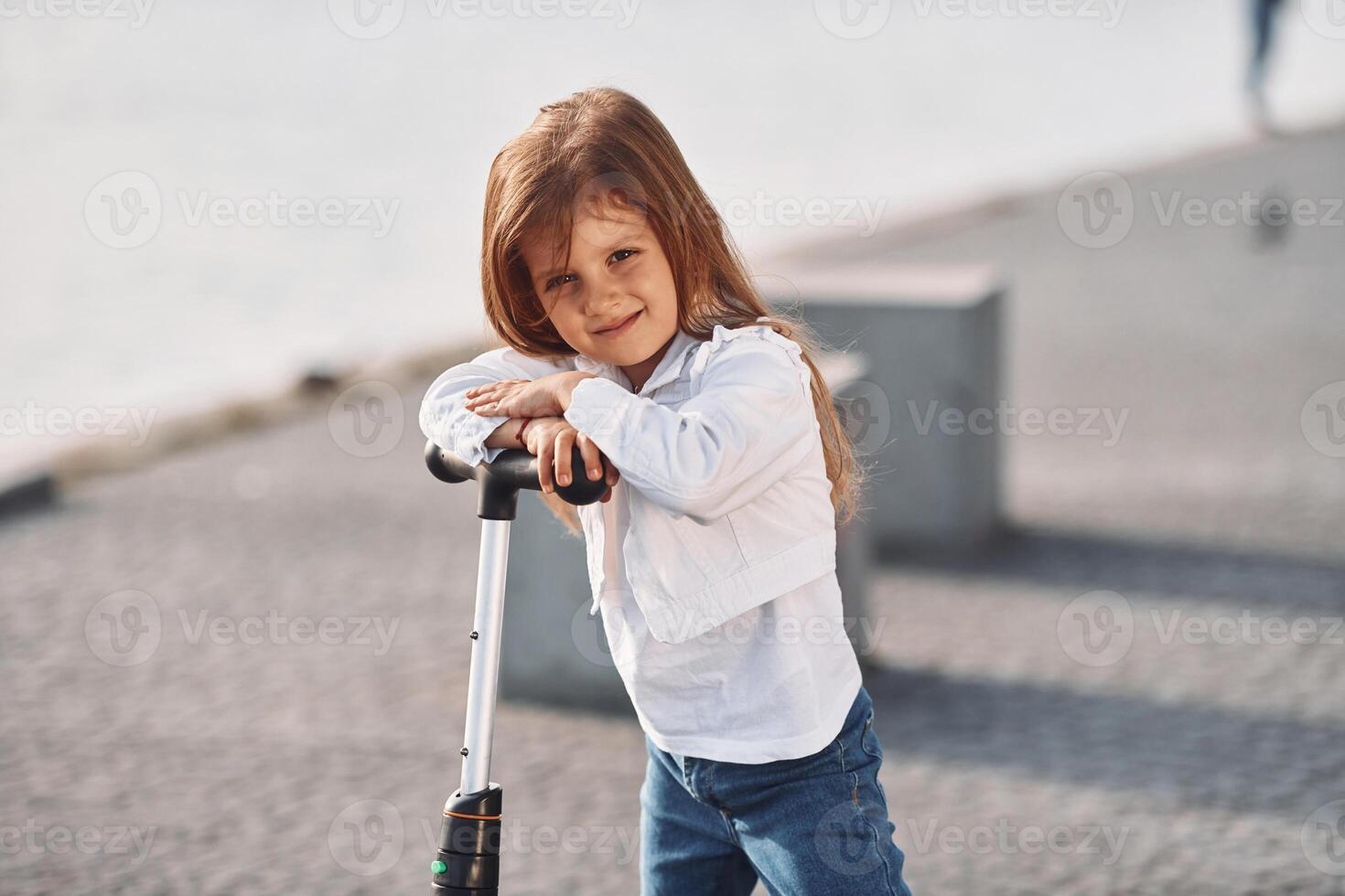Standing and looking into the camera. Cute little girl with scooter is outdoors in summertime photo