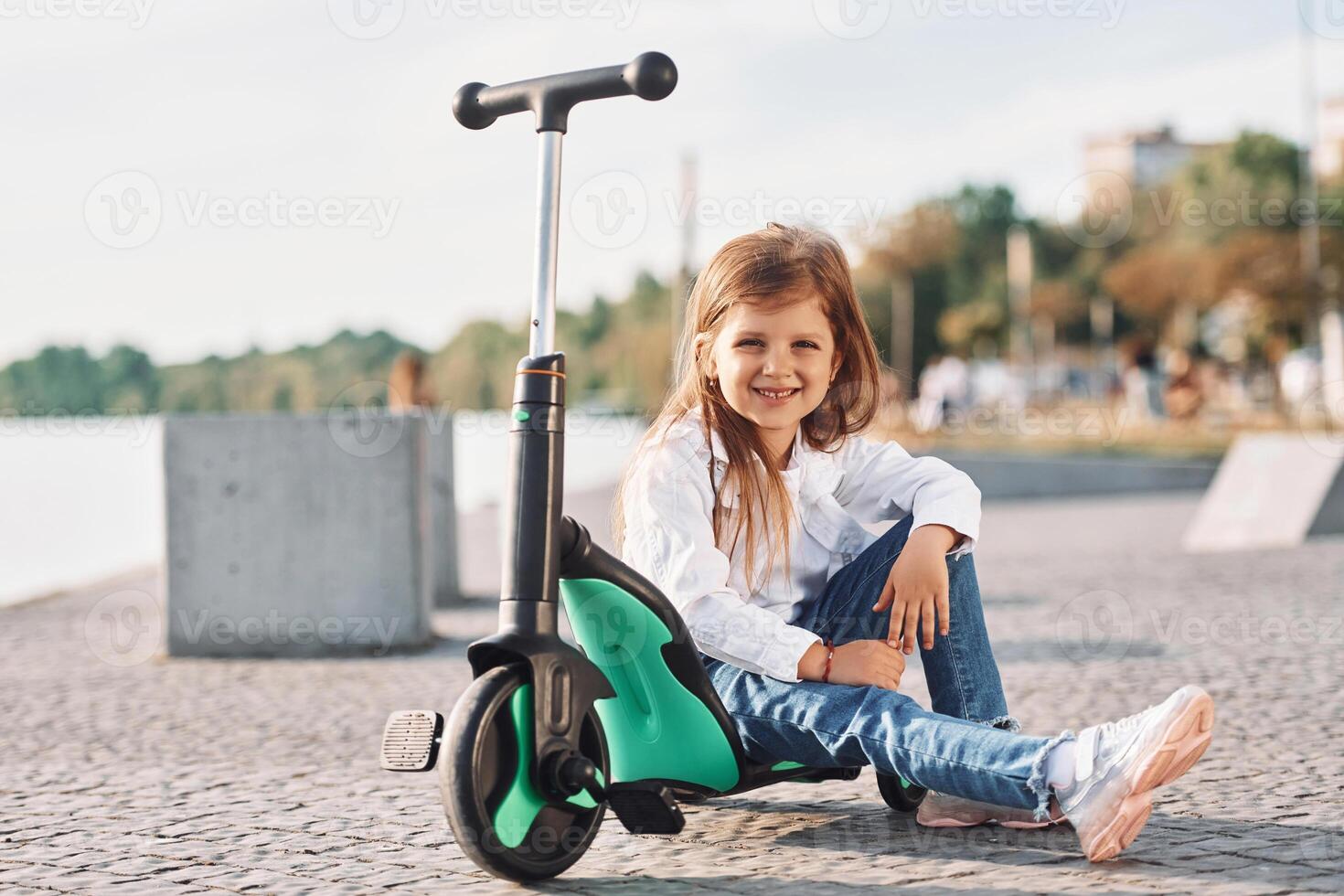 Sitting and smiling. Cute little girl with scooter is outdoors in summertime, photo