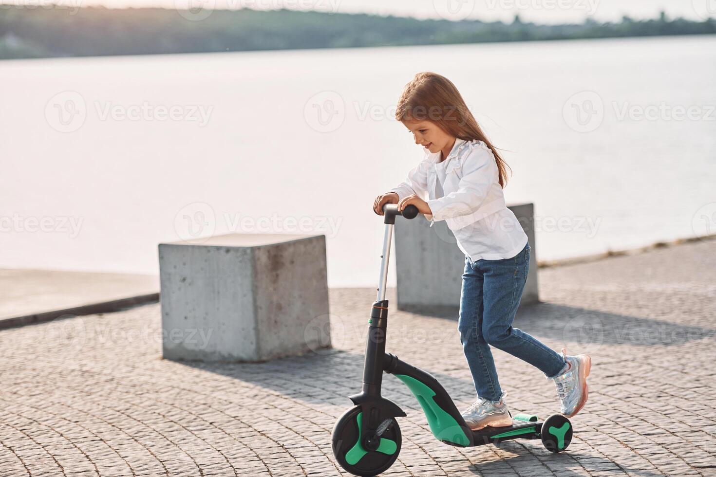 Riding the vehicle. Cute little girl with scooter is outdoors in summertime photo