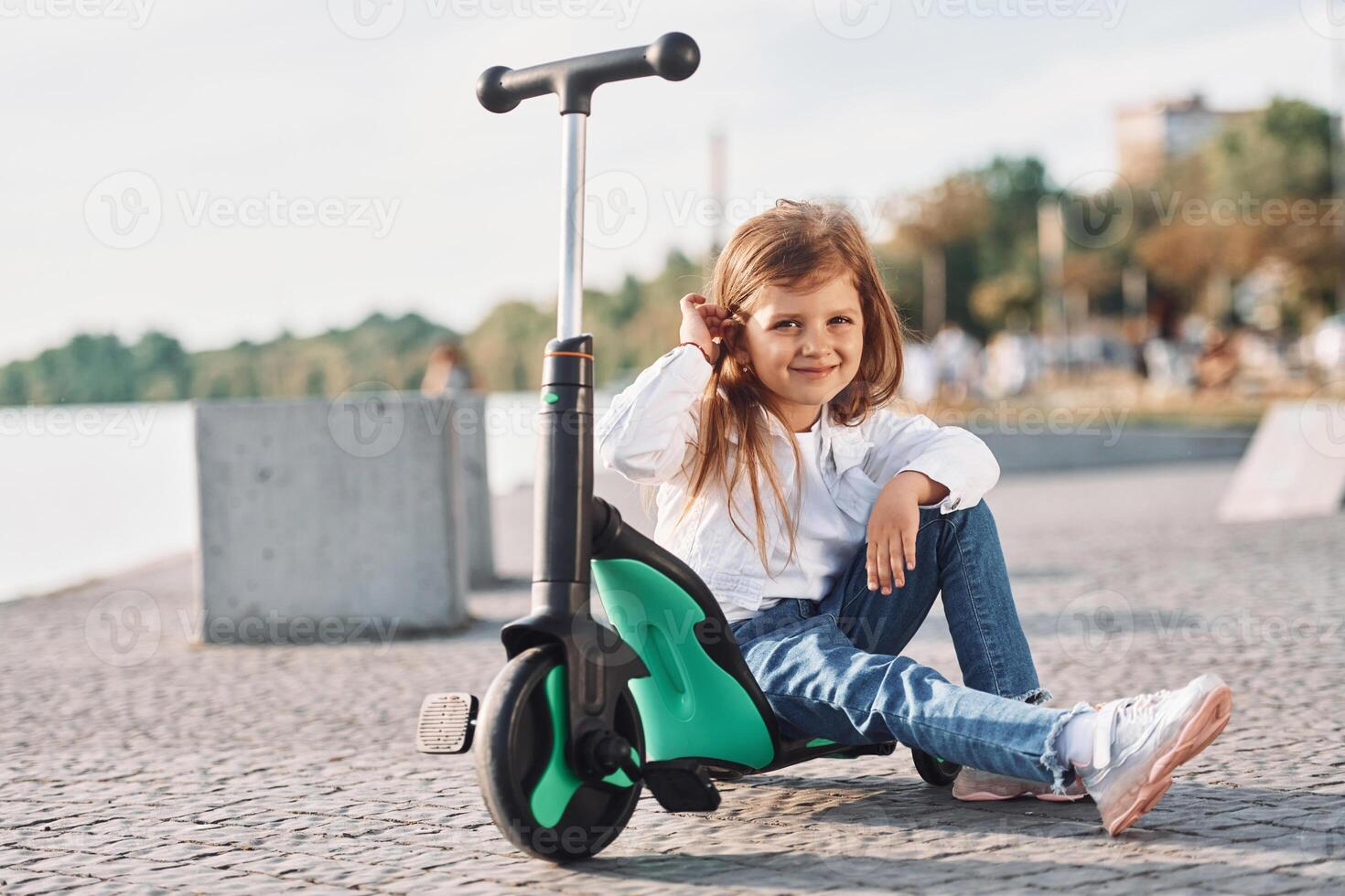 Sitting and smiling. Cute little girl with scooter is outdoors in summertime,. photo