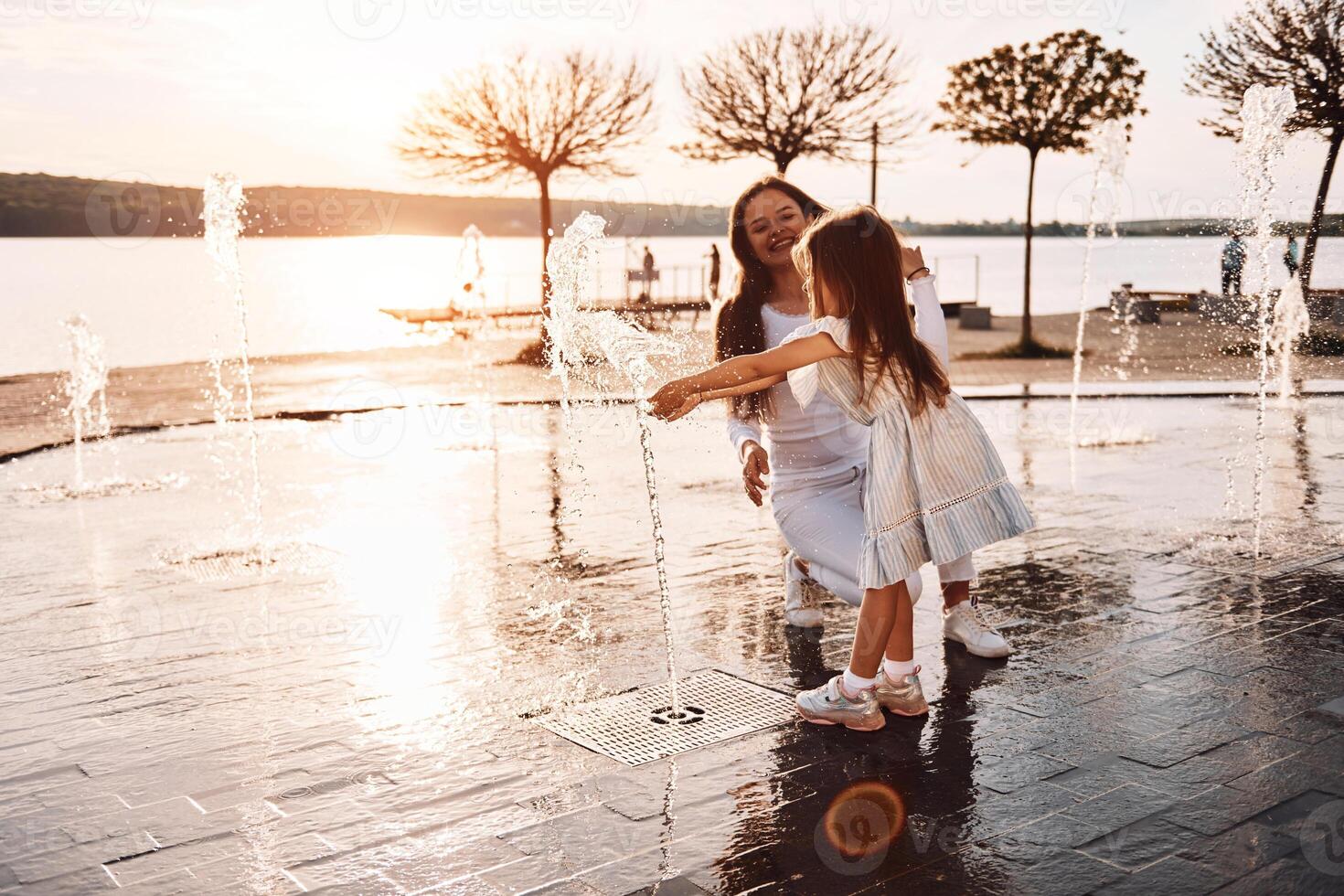 Holding each other. Little girl with her mother having fun outdoors in fountain near the lake, photo