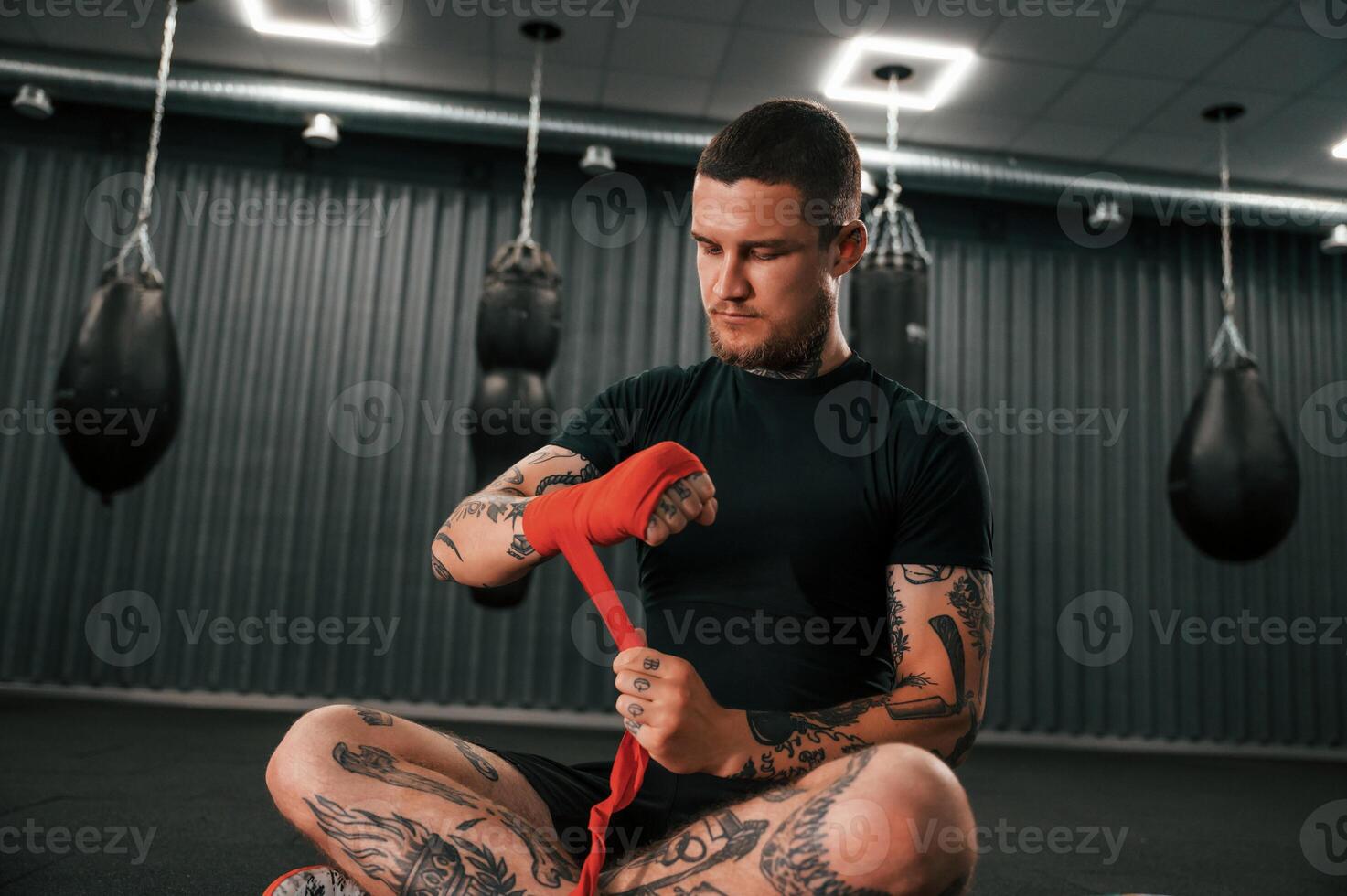 Sitting and wearing red colored hand wraps. Young tattooed man is practicing boxing indoors in the gym. photo