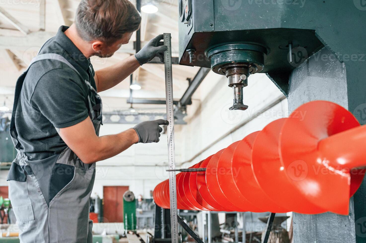 Measuring red colored sharp part. Man in uniform is in workstation developing details of agriculture technique., photo