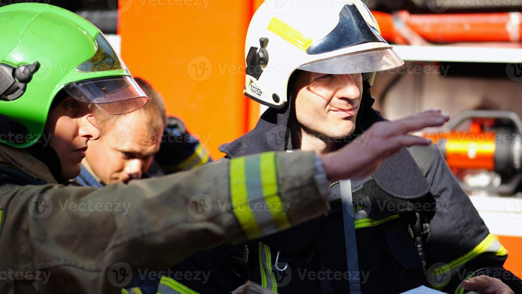 Male firefighters in protective uniforms discuss rescue plan looking at paper instructions near fire truck. Young firemen prepare to extinguishing fire against the background of big red car. Slow mo photo