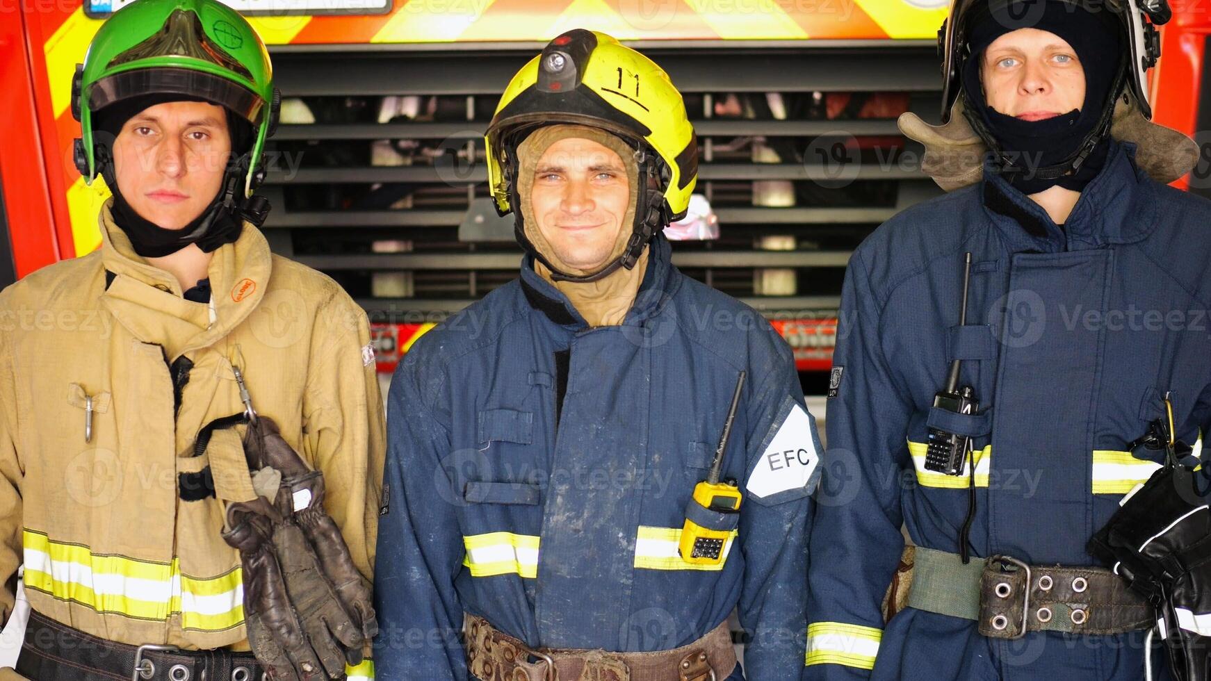 Portrait of young happy fireguards in protective uniforms against background of fire truck at station. Male firefighters in full equipment looking into camera with positive emotions near big red car photo