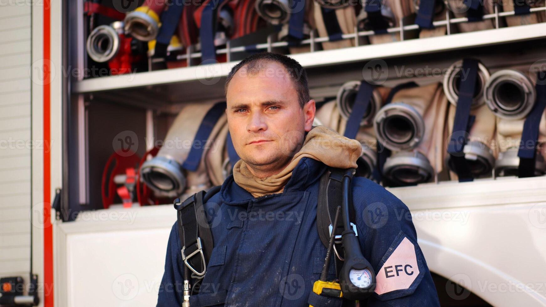 Portrait of confident male firefighter standing near a fire engine. Young fireman in protective uniform posing against the background of big red truck. Concept of saving lives and heroic profession photo
