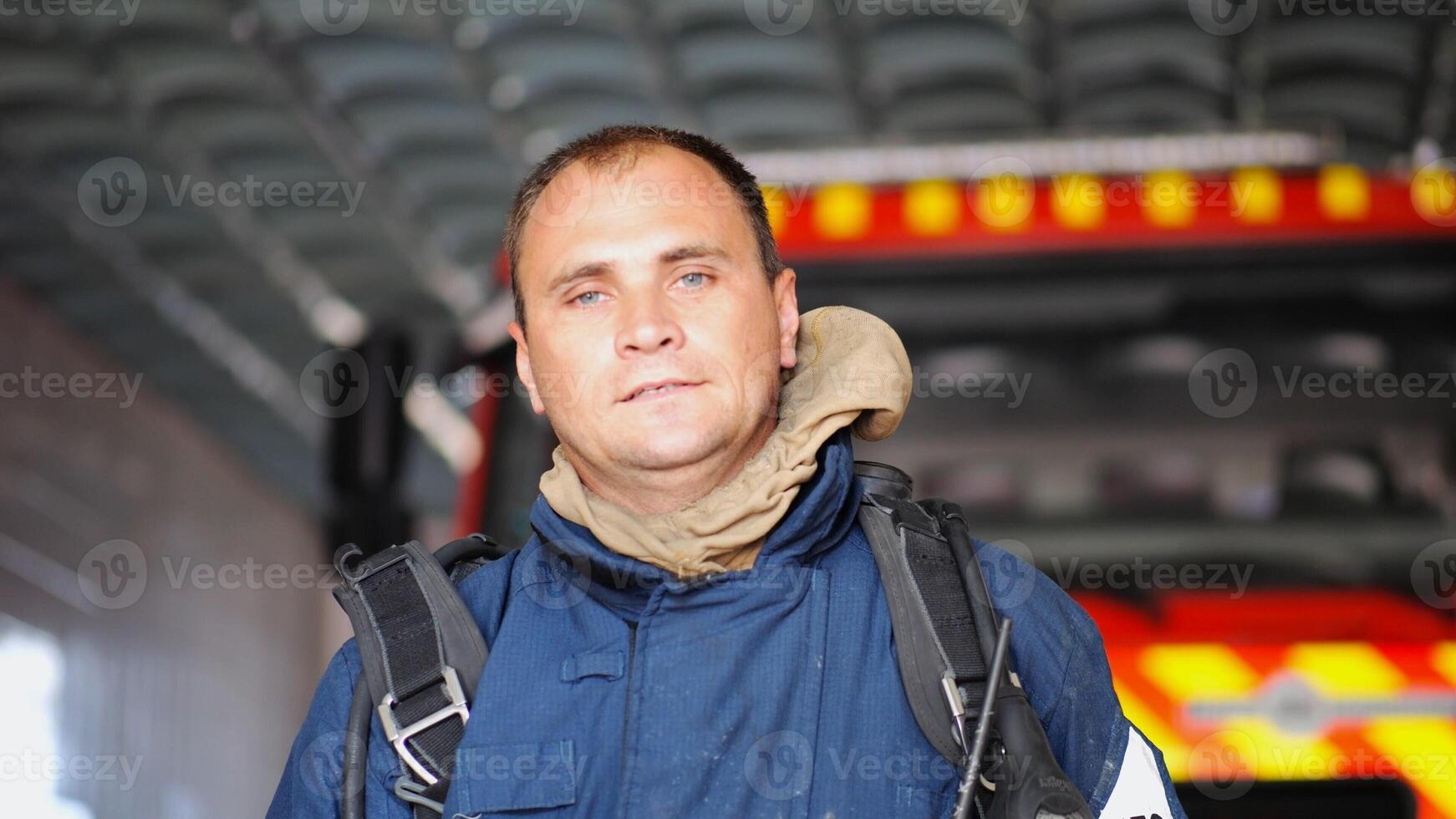 Portrait of tired male firefighter standing near a fire engine at department. Young exhausted fireman in protective uniform looking at camera after fire against the background of big red truck photo