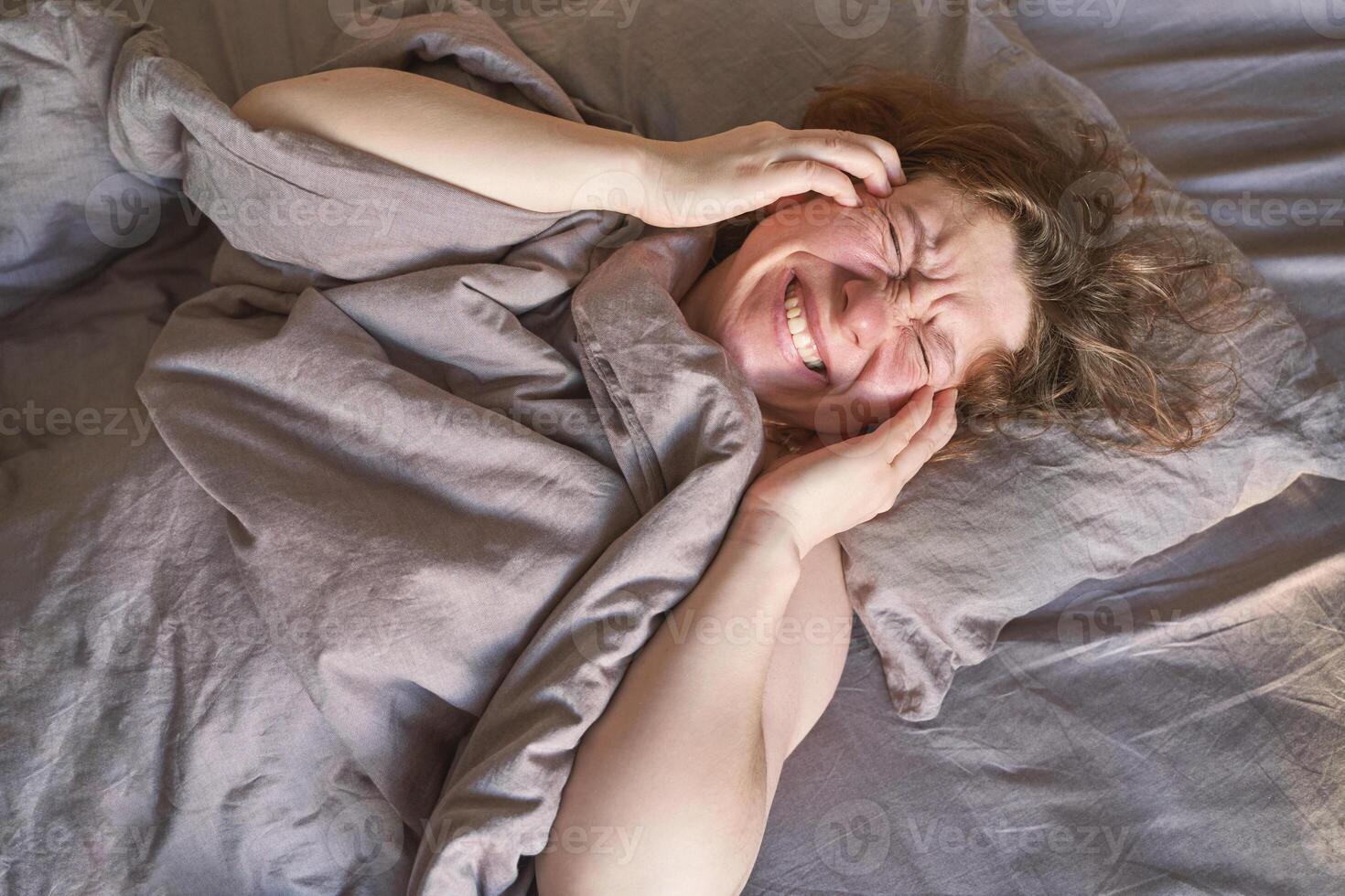 Mature caucasian female joyfully lying in bed under gray sheets smiling with eyes closed. photo