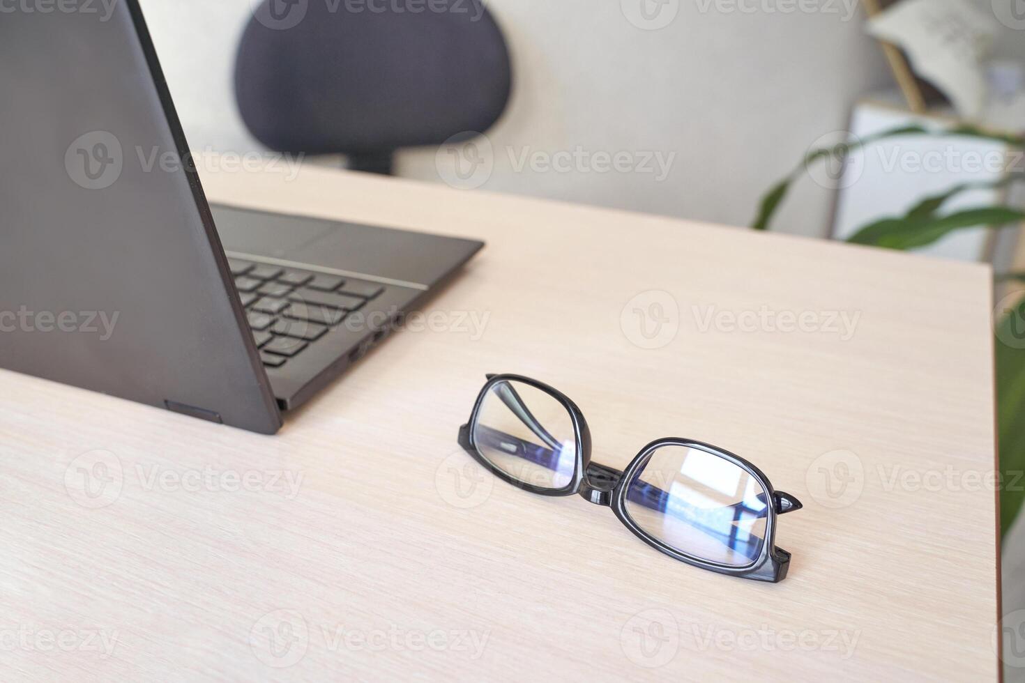 Eyeglasses resting on a wooden desk beside open laptop in modern workspace with black chair. photo