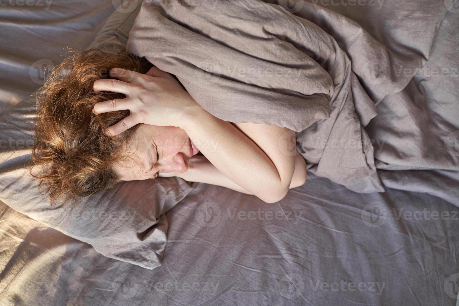 Caucasian female adult experiencing stress or anxiety while lying in bed under gray blanket on a bright morning. photo