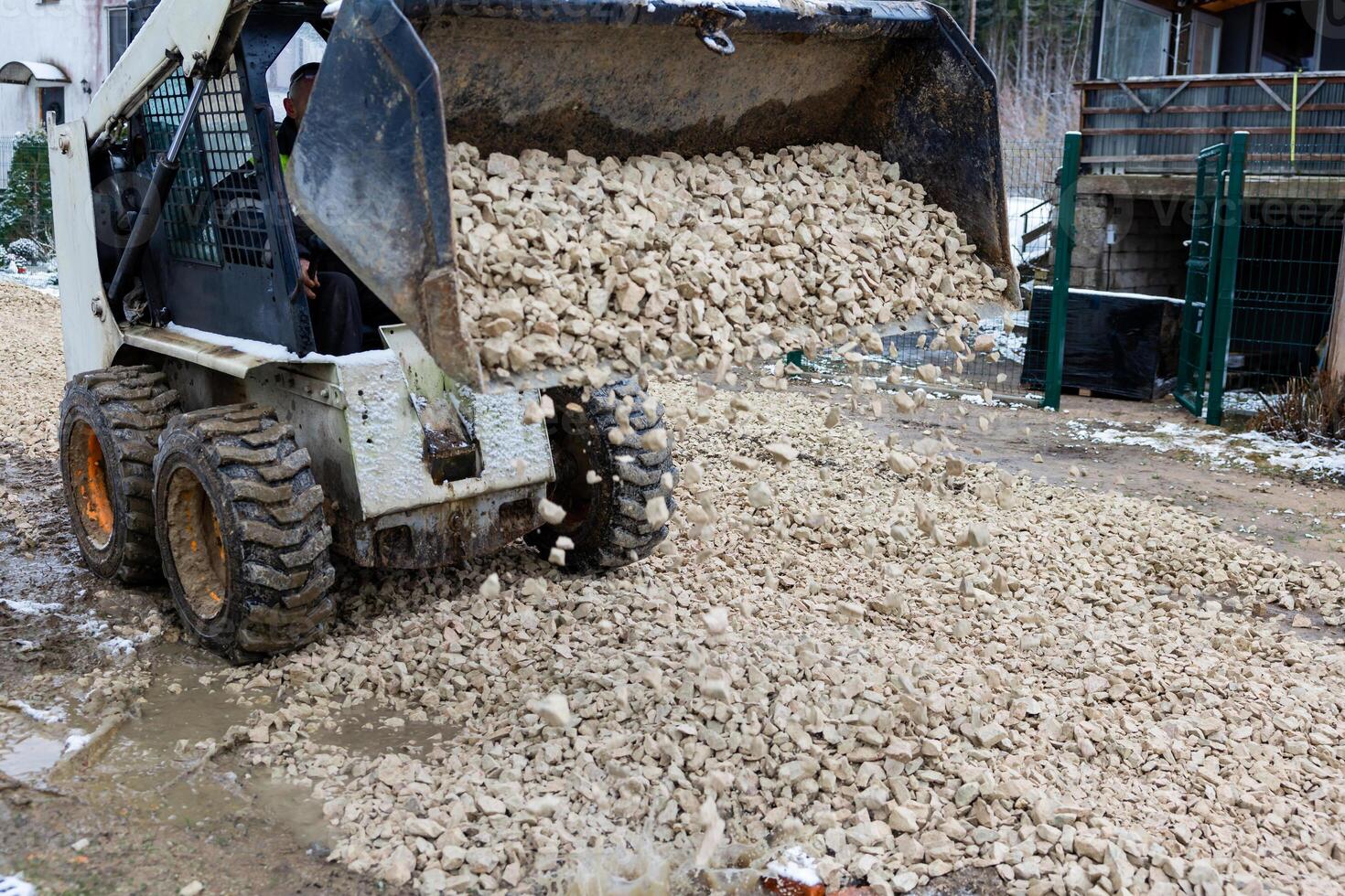 Skid Steer Loader Dumping Crushed Stones on Muddy Ground in Overcast Weather photo