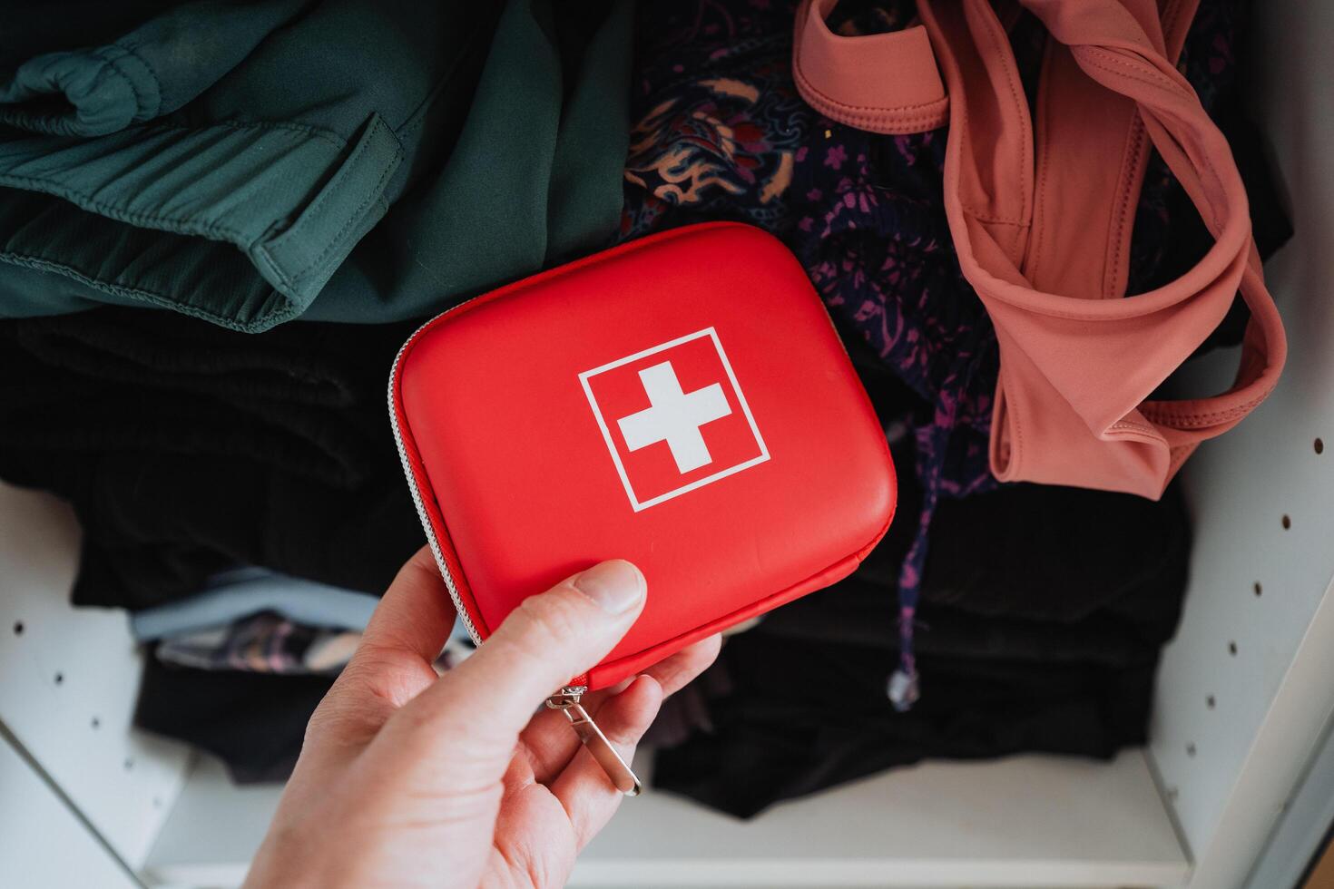 Compact Red First Aid Kit Held by Hand Amongst Clothing in a Neatly Organized Closet photo