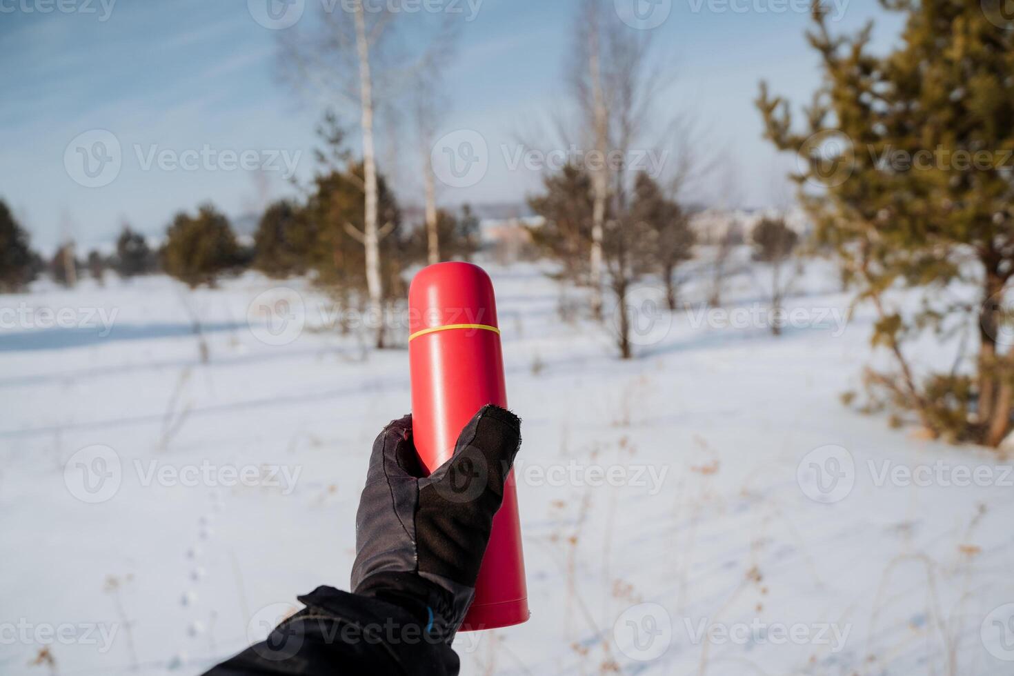 A person is bundled up in warm and cozy winter clothing, standing in the midst of pristine white snow, while holding a bright red thermos that starkly contrasts with the icy white backdrop photo