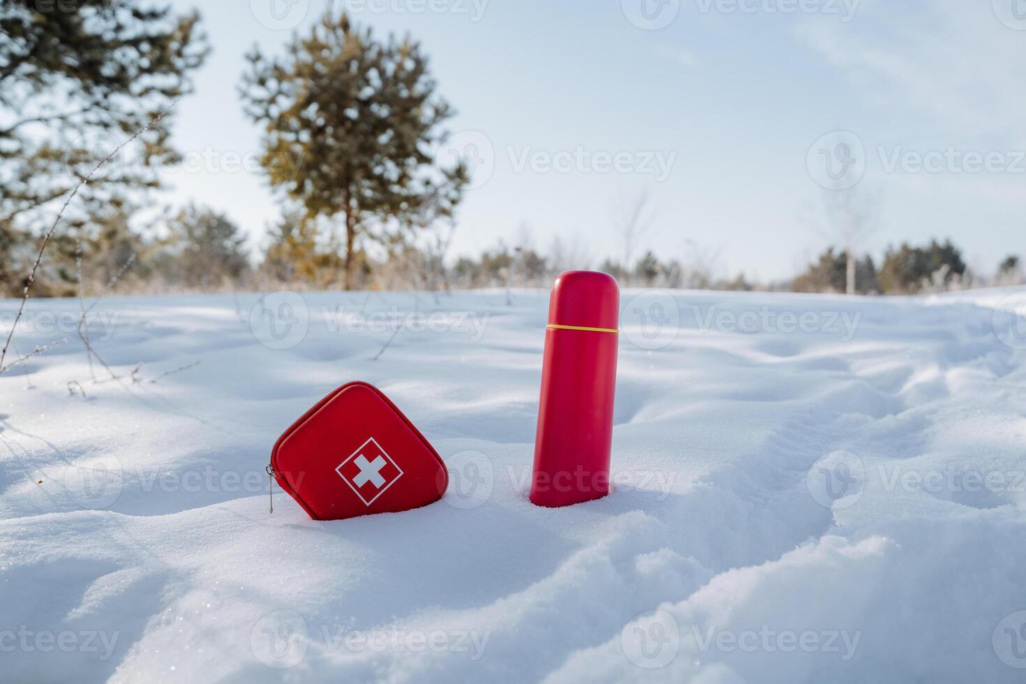 In the thick, soft layers of snow, you can spot a bright red first aid kit alongside a matching red thermos, both standing out distinctly against the white, icy landscape photo