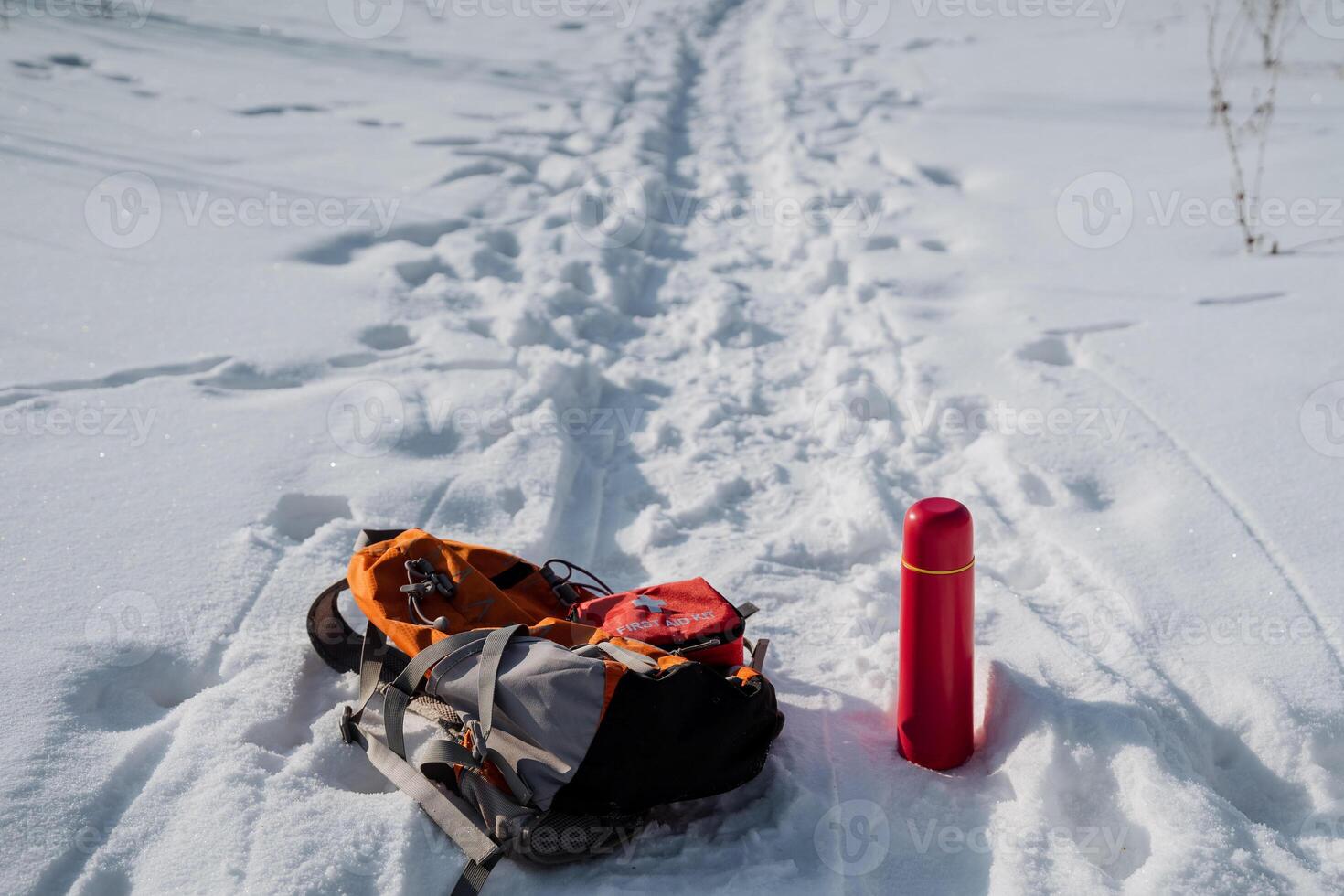 A colorful backpack, paired with a bright red thermos, is resting in the pristine white snow, producing a striking contrast to the beautiful winter landscape surrounding it all photo