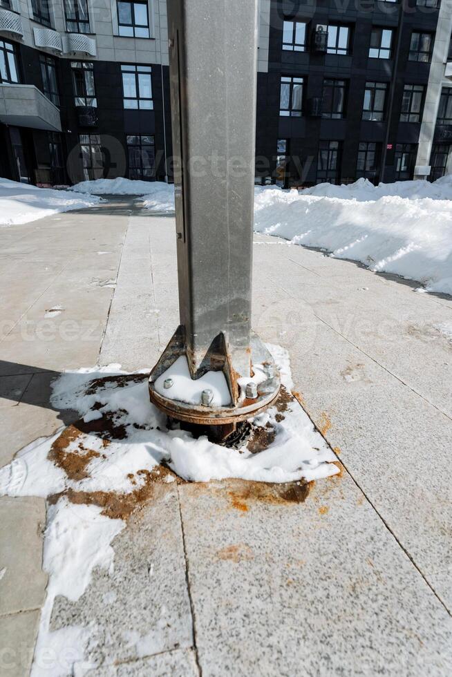 In an urban setting, there is a rusty base of a metal pole that is surrounded by a blanket of snow, showcasing a stark contrast between decay and the pristine winter landscape photo