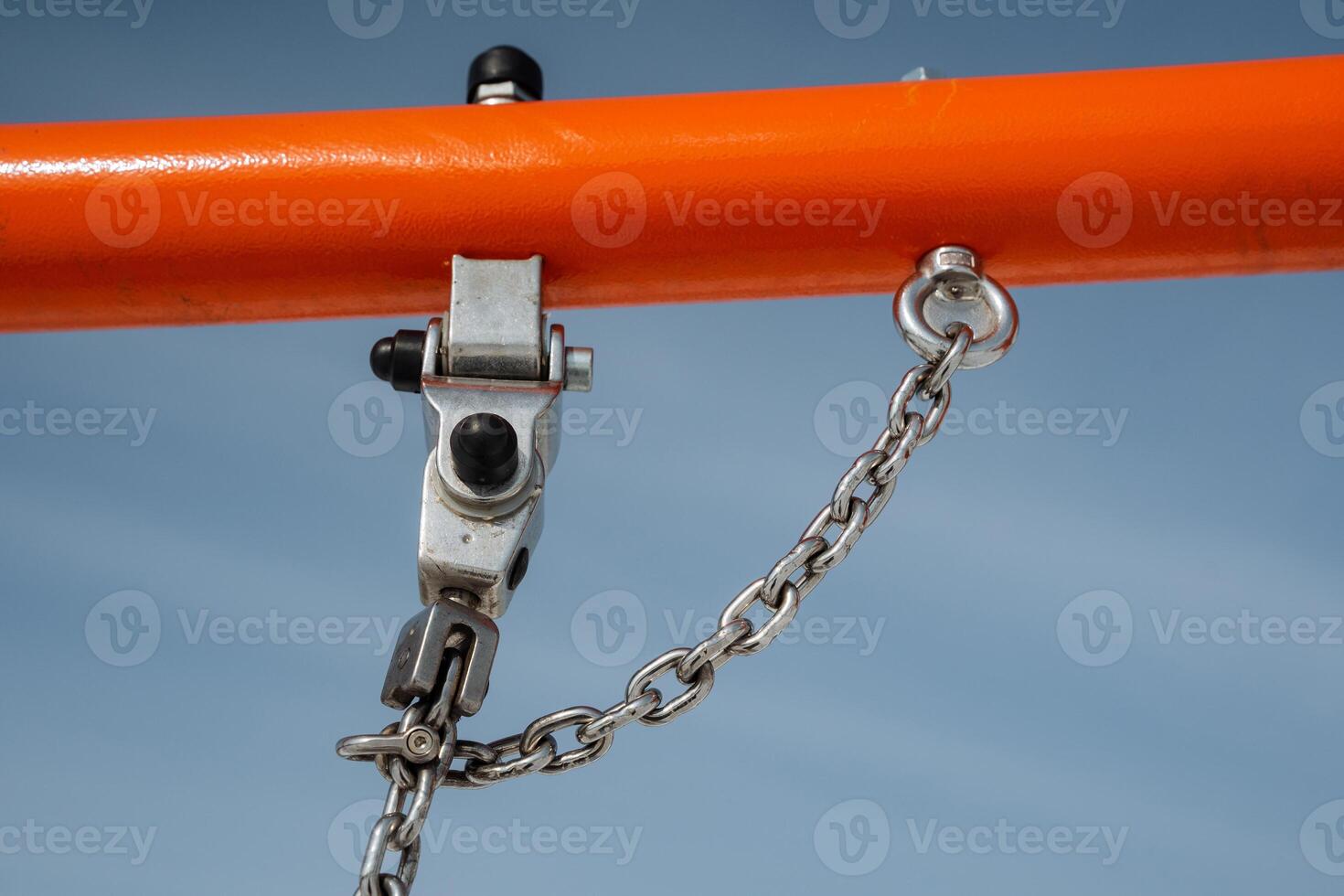 A detailed closeup view featuring a sturdy metal hook and robust chain mounted on a vibrant orange frame, set against a backdrop of a clear blue sky, showcasing industrial design photo