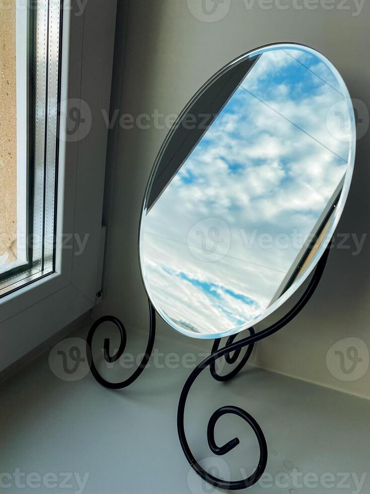 A beautifully crafted round mirror, made with exquisite materials, is placed on a window sill, reflecting the soft, warm natural light streaming in from the charming window photo