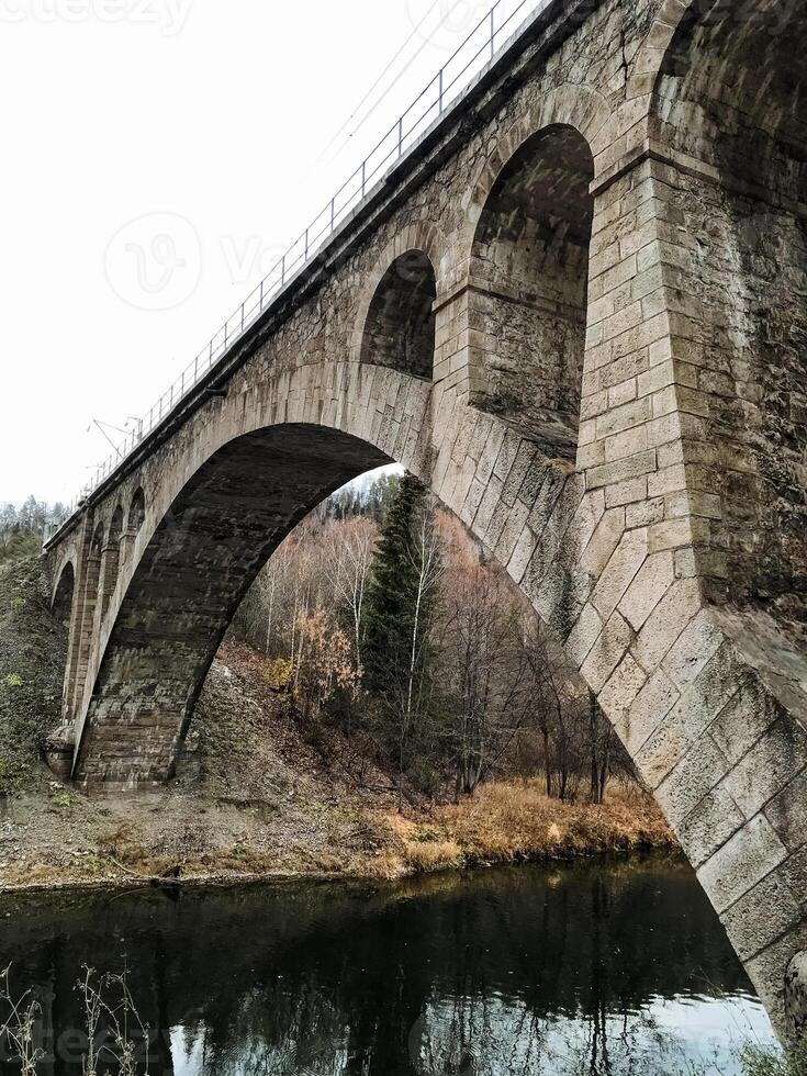 A beautiful stone bridge arches gracefully over a flowing river, nestled in the heart of a lush, vibrant forest, creating a picturesque scene that captures the natural beauty around it photo