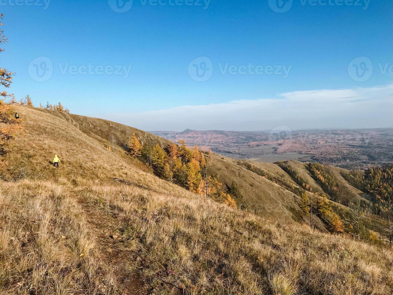 A person confidently stands on a lush green hill, surrounded by breathtaking mountains and a vast wilderness that stretches into the horizon, showcasing stunning natural beauty photo