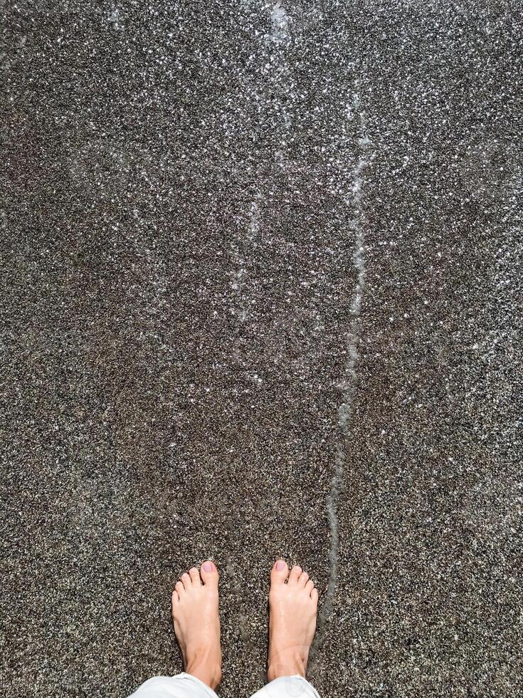 A persons feet are standing on a wet concrete surface that has recently been poured, showcasing the texture and dark sheen of the freshly mixed concrete underneath them photo