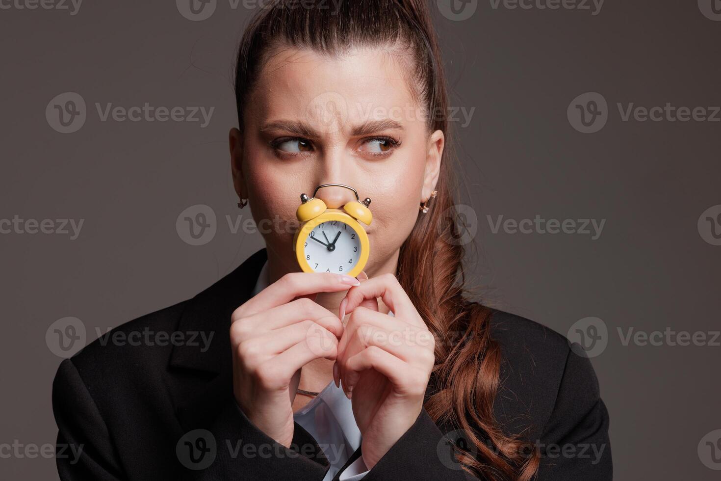 Businesswoman holding a small yellow alarm clock near her face with a skeptical expression, symbolizing doubt, time pressure, and decision-making photo