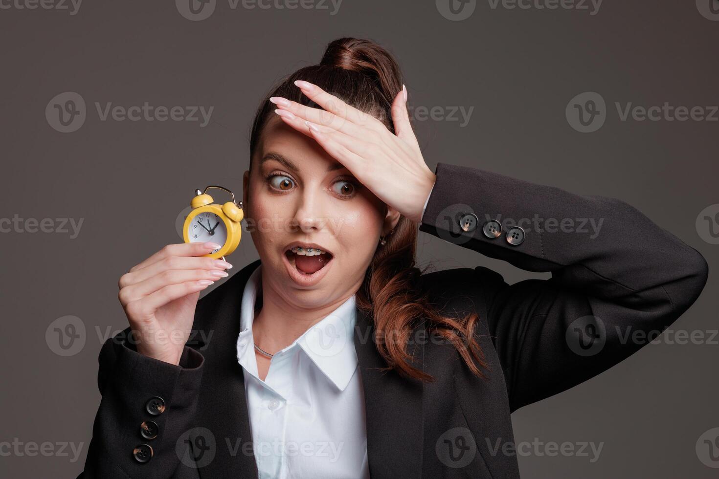 Stressed businesswoman in a suit holding a small alarm clock, shocked at the time, running late, concept of time management and deadline pressure photo