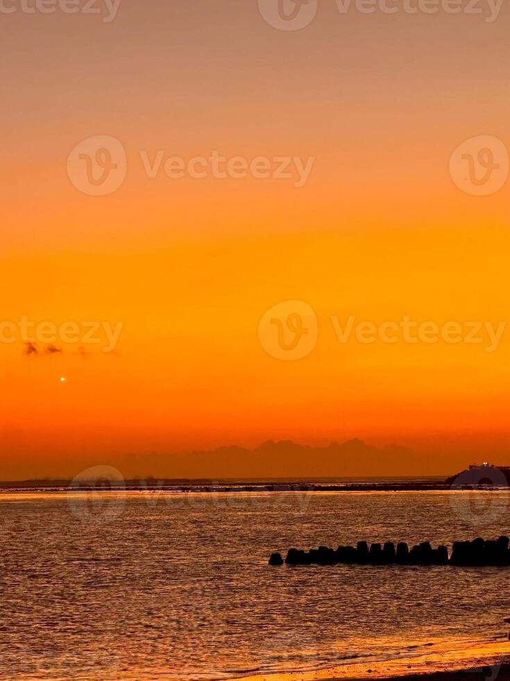 Orange sunset over Kelan Beach, Bali, with calm ocean waves and a scenic breakwater. A tranquil coastal scene with warm hues and serene atmosphere. photo