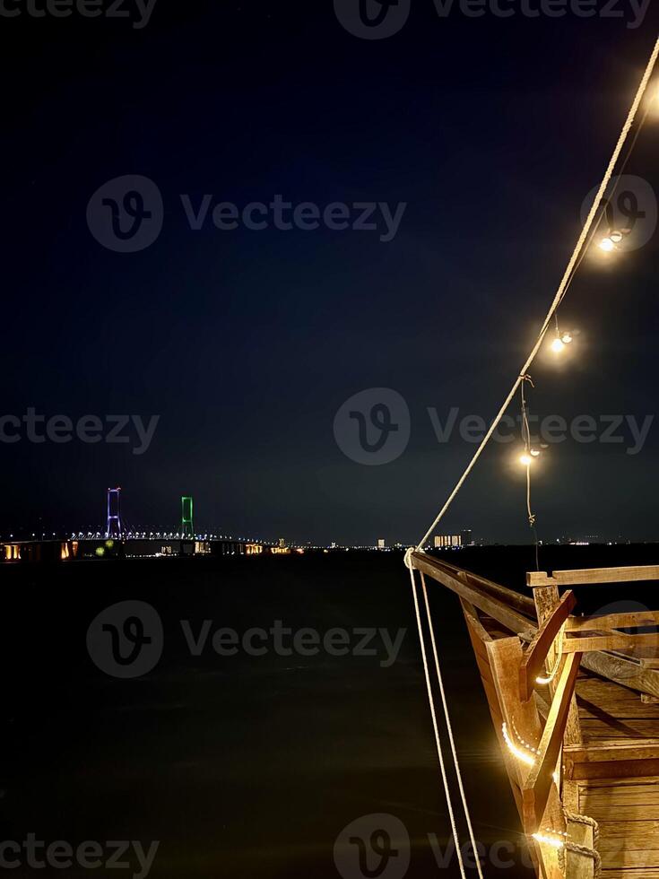 Rustic Ship-Themed Cafe with a Wooden Helm Overlooking the Madura Strait and Suramadu Bridge. Unique Boat Deck Ambiance, Nautical-Style Outdoor Dining, Coastal View with Sunset Sky. photo