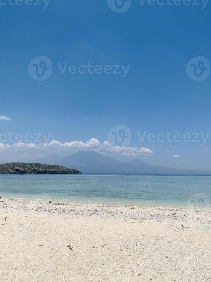 Menjangan Island, West Bali, with crystal-clear blue waters and pristine white sands. Three majestic mountains in the distance frame the panorama, creating a serene natural atmosphere. photo