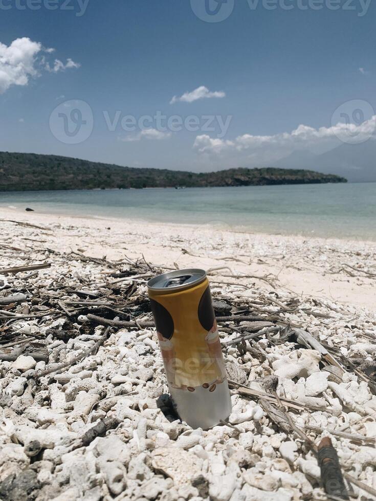 A close-up of a rebranded beverage can rests on the beach, its sleek design contrasts with rugged rocks, branches, and a vast blue ocean. Distant hills add natural calm, pure bliss. photo
