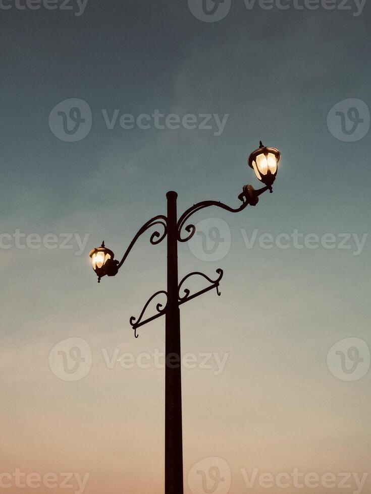 A vintage street lamp from a low angle warm light contrasts a sky shifting from dusk blue to soft orange. Twilight's nostalgic charm crowns this serene sentinel. photo