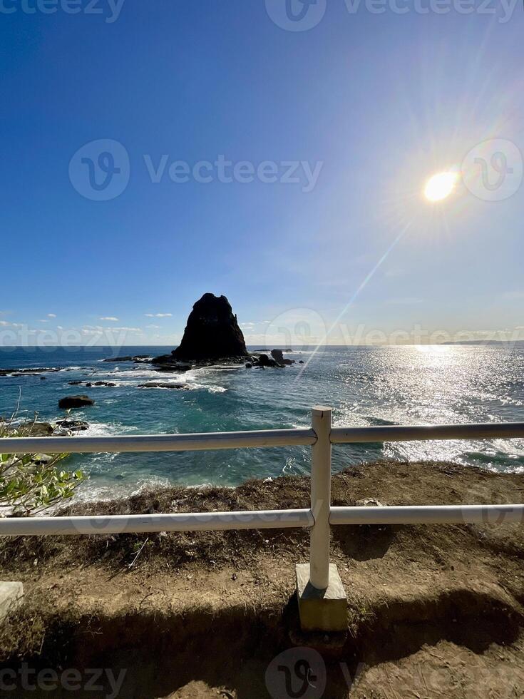 A breathtaking view of Papuma Beach at midday from an elevated vantage. The deep blue ocean meets a clear sky as a rugged rocky islet rises, framed by a wooden fence. A dream view. photo