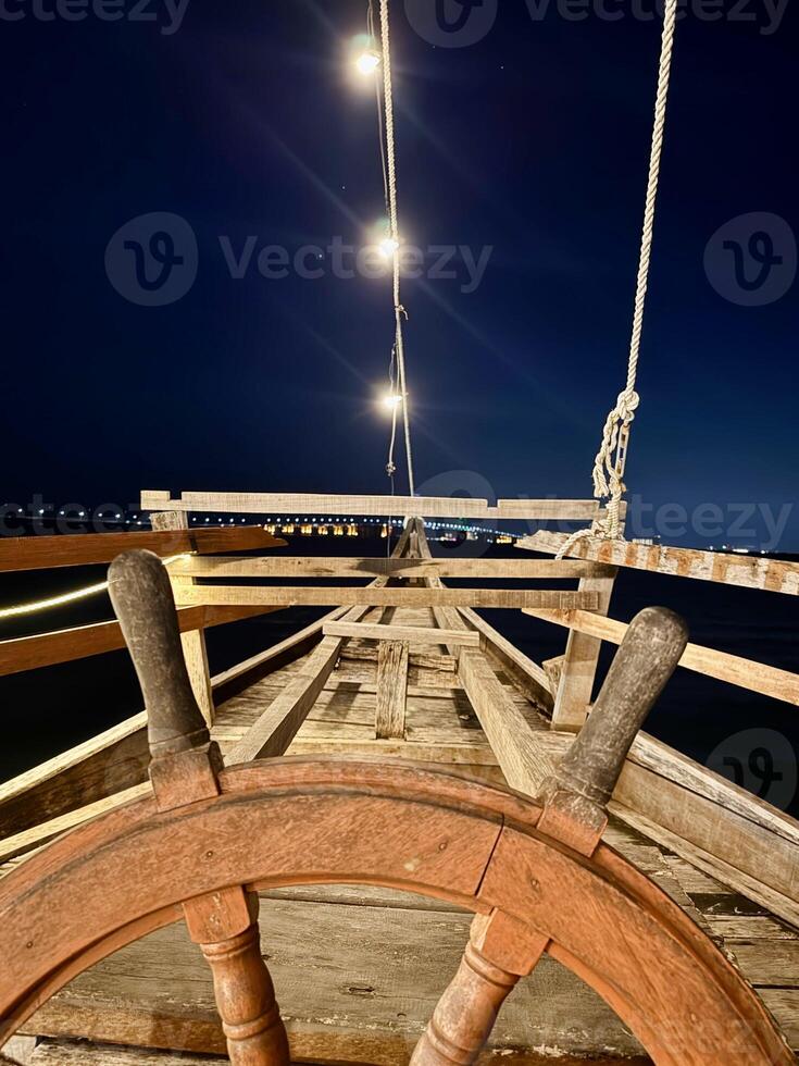Rustic Ship Themed Cafe with a Wooden Helm Overlooking the Madura Strait and Suramadu Bridge. Unique Boat Deck Ambiance, Nautical-Style Outdoor Dining, Coastal View with Sunset Sky. photo