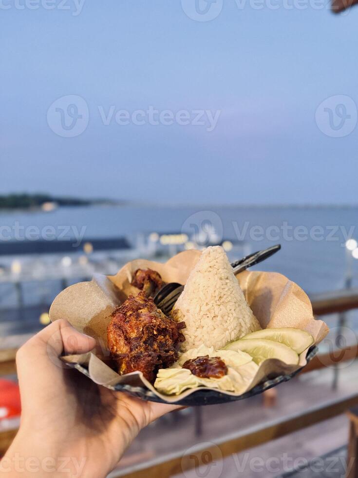 Indonesian Fried Chicken with Rice and Sambal, Served with Fresh Vegetables Overlooking the Ocean. Traditional Nasi Ayam, Street Food, Dining, Scenic View, Culinary Travel, Southeast Asian Cuisine. photo