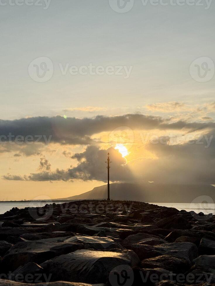 Serene Sunrise Over Rocky Jetty with a tall pole silhouetted with overcast cloud. Plengsengan Beach Banyuwangi, Bali Strait photo