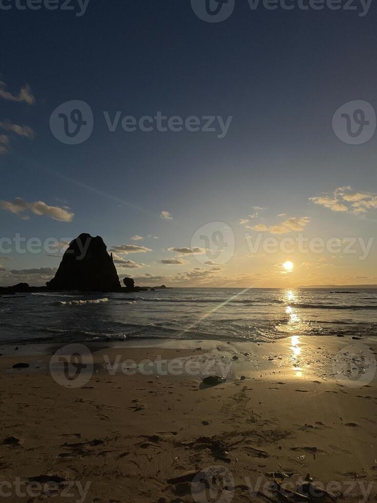 landscape view of Papuma Beach in Jember, featuring a rocky islet standing tall against the warm hues of the setting sun. The sky transitions from deep blue to orange. Papuma Beach Jember photo