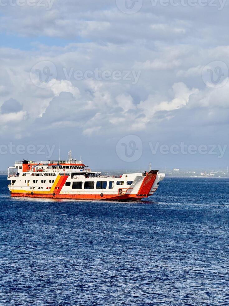 Large ferry sailing across the Bali Strait under a cloudy sky with deep blue ocean water. Passenger and vehicle transport ferry connecting Bali and Java, Indonesia, with open ramp and vibrant colors. photo