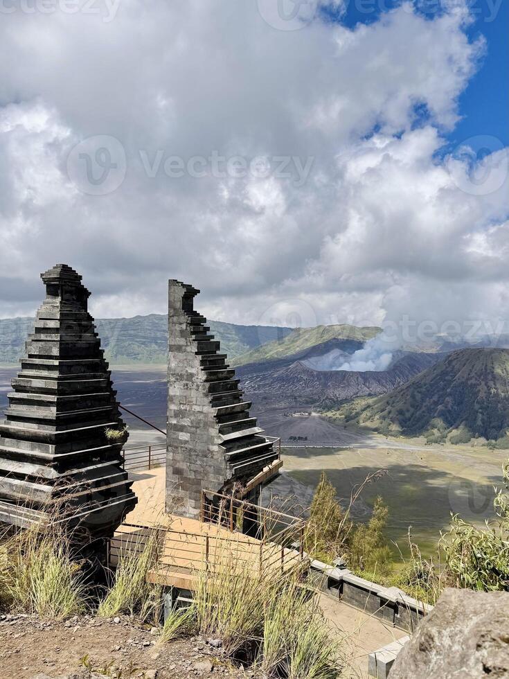 View of Mount Bromo with traditional Balinese split gate in the foreground under a blue sky with clouds. Stunning volcanic landscape, lush green hills, and volcanic smoke in East Java, Indonesia. photo