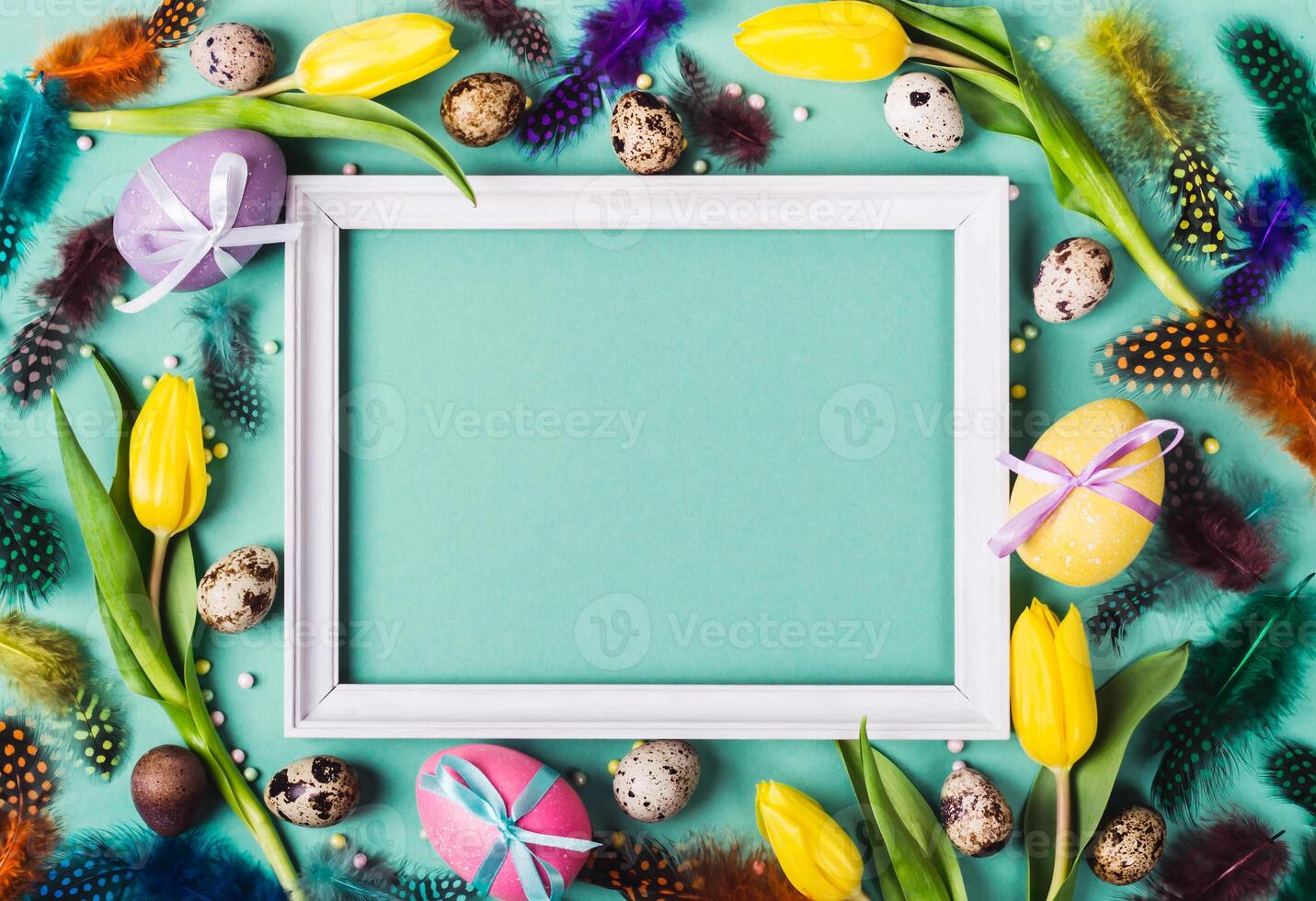 Easter Frame of yellow tulips, decorated eggs and feathers on turquoise background for greeting message photo