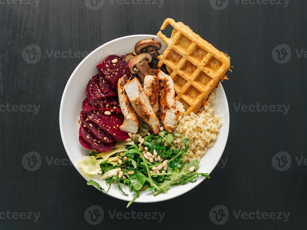 Healthy Food concept. Bowl with chicken, mushrooms, beetroot, quinoa, green rucola, decorated pine nuts and carrot waffle. Top view of gourmet bowl. Balanced gourmet dish on black wooden background photo