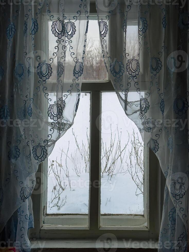 A rustic wooden window with delicate lace curtains in a grandmother's house in a Russian village. The embroidered fabric softly filters the daylight, revealing a serene winter landscape outside. Snow photo