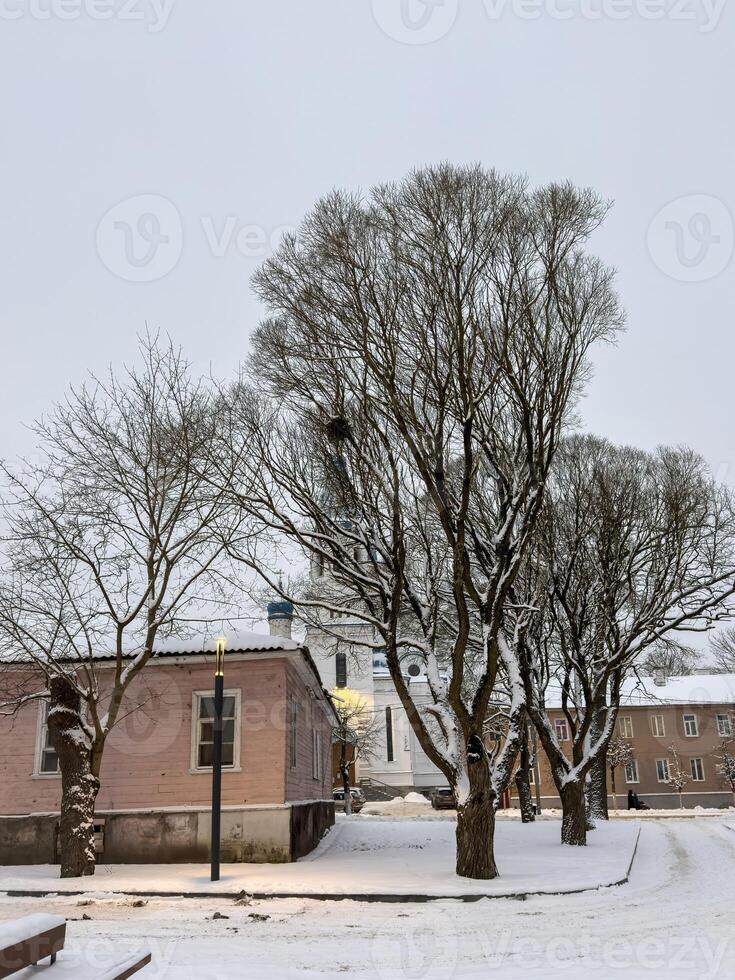 A quiet winter street scene featuring snow-covered trees, a small wooden house, and a distant church with blue domes. The cold atmosphere and evening light create a peaceful and nostalgic setting photo