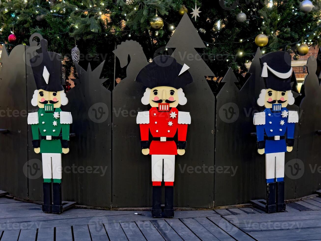 Three wooden nutcracker soldiers in red, green, and blue stand in front of a beautifully decorated Christmas tree. Their traditional military-style uniforms and cheerful expressions add a festive photo